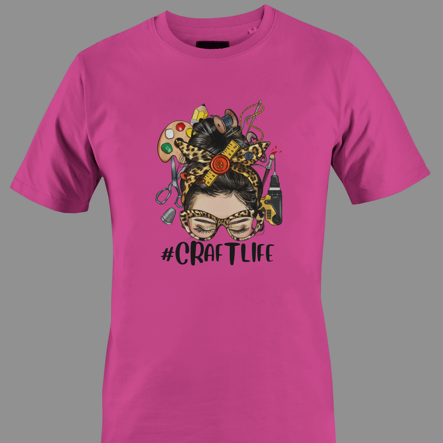Dark pink t-shirt with a graphic design featuring a woman's face with craft supplies around her head, featuring the hashtag #CRAFTLIFE.
