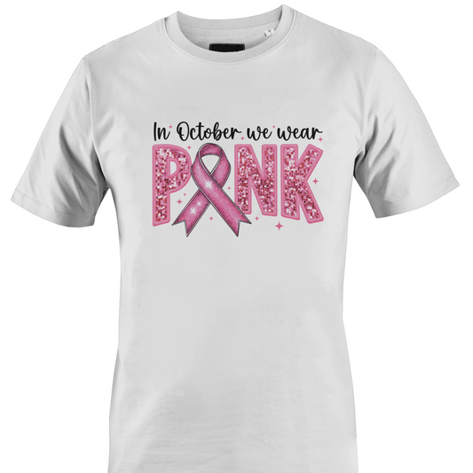 A white short-sleeve crewneck t-shirt featuring a pink graphic on the front that reads 'In October we wear PINK' with a pink ribbon and glitter-textured letters by Monkey's Creations. 