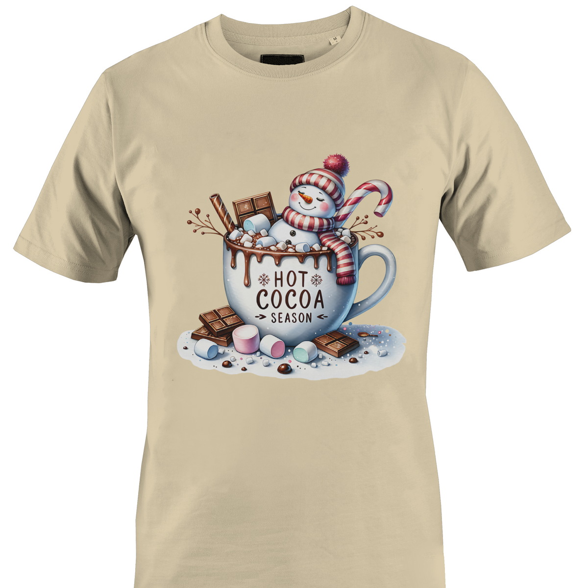 A beige short-sleeve crewneck t-shirt featuring a colorful graphic print of a winter snowman wearing a red and white striped scarf, sitting in a white mug filled with hot cocoa, chocolate chunks, marshmallows, and a candy cane. The text 'HOT COCOA SEASON' is printed on the mug by Monkey's Creations.