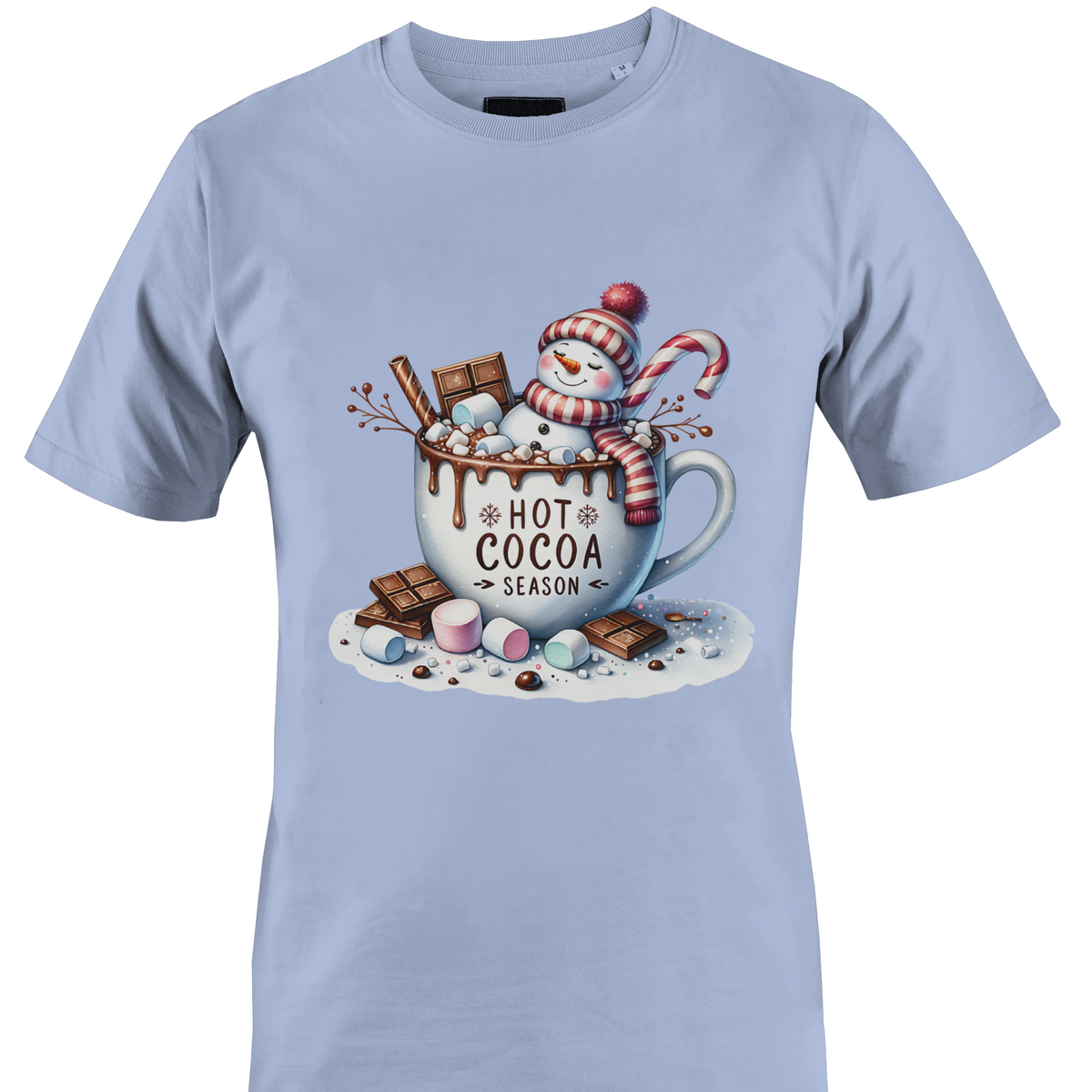 A light blue short-sleeve crewneck t-shirt featuring a colorful graphic print of a winter snowman wearing a red and white striped scarf, sitting in a white mug filled with hot cocoa, chocolate chunks, marshmallows, and a candy cane. The text 'HOT COCOA SEASON' is printed on the mug by Monkey's Creations.
