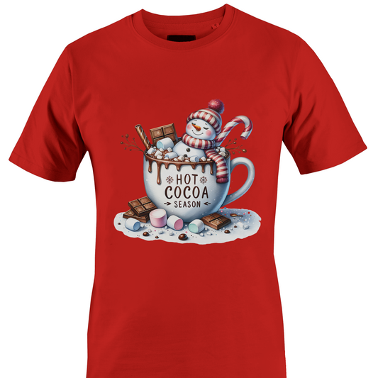 A red short-sleeve crewneck t-shirt featuring a colorful graphic print of a winter snowman wearing a red and white striped scarf, sitting in a white mug filled with hot cocoa, chocolate chunks, marshmallows, and a candy cane. The text 'HOT COCOA SEASON' is printed on the mug by Monkey's Creations.