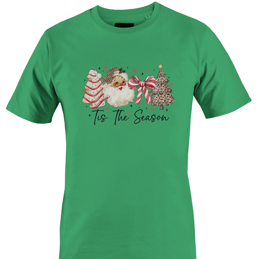 A green short-sleeve crewneck t-shirt featuring a festive Christmas graphic on the chest. The graphic depicts a watercolor Santa Claus, a decorated Christmas tree, a striped ornament, and a large red bow, with the text 'Tis The Season' written below in a black script font by Monkey's Creations.