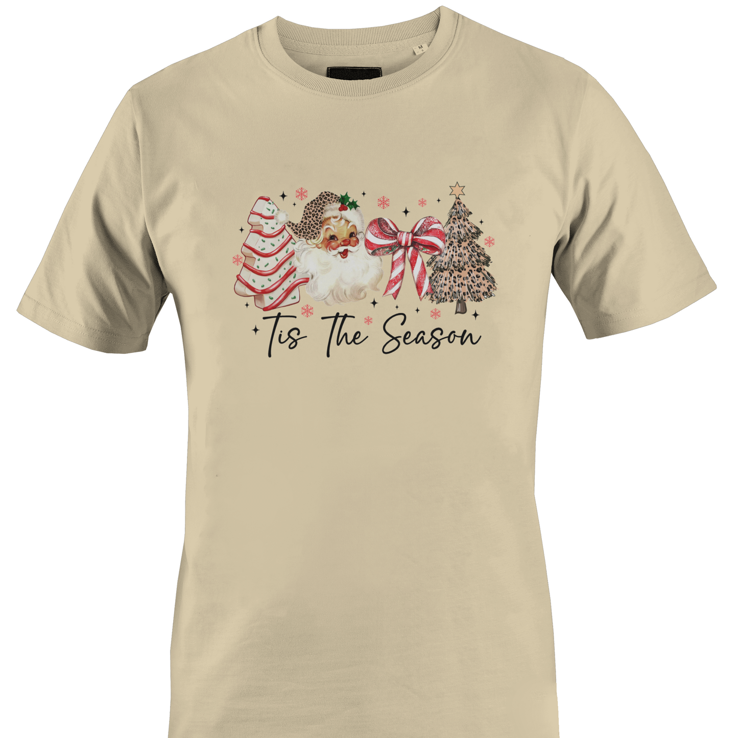 A tan short-sleeve crewneck t-shirt featuring a festive Christmas graphic on the chest. The graphic depicts a watercolor Santa Claus, a decorated Christmas tree, a striped ornament, and a large red bow, with the text 'Tis The Season' written below in a black script font by Monkey's Creations.